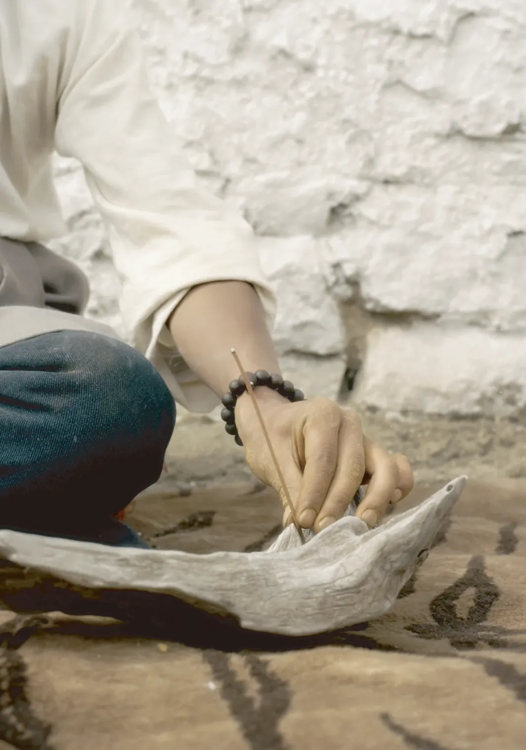 A hand setting a lit incense stick into a weathered driftwood holder beside a whitewashed wall.