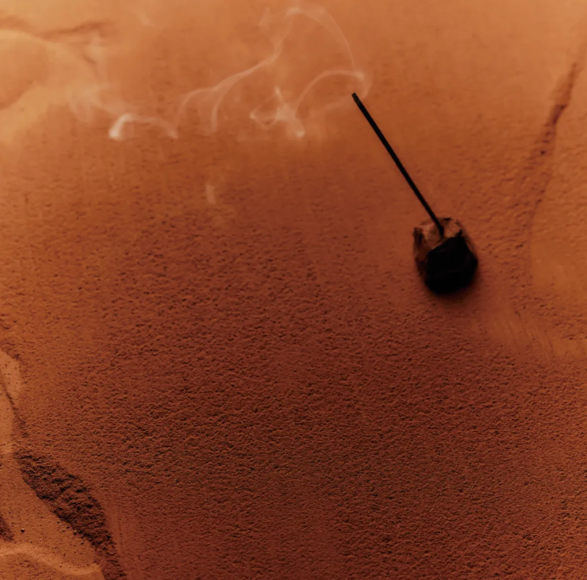 A single incense stick smoldering in a small holder, a thread of smoke drifting across a terracotta surface.